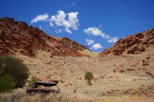 An old rusty car sits at the base of a red rocky range, in a remote landscape.